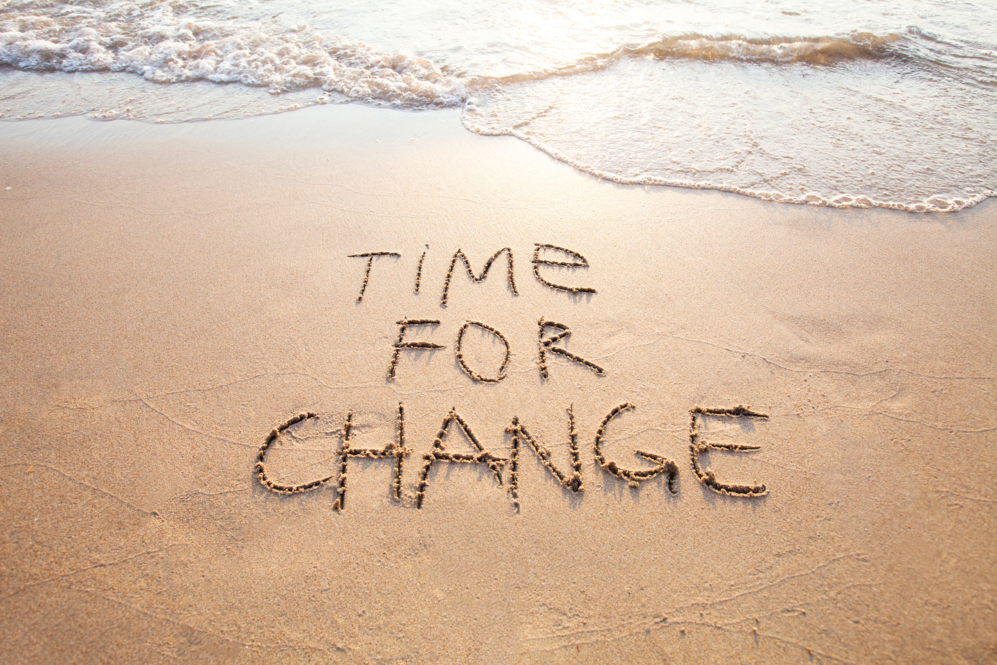 Die Worte "TIME FOR CHANGE" sind im Sand am Strand geschrieben, während Wellen im Hintergrund sanft ans Ufer schlagen.